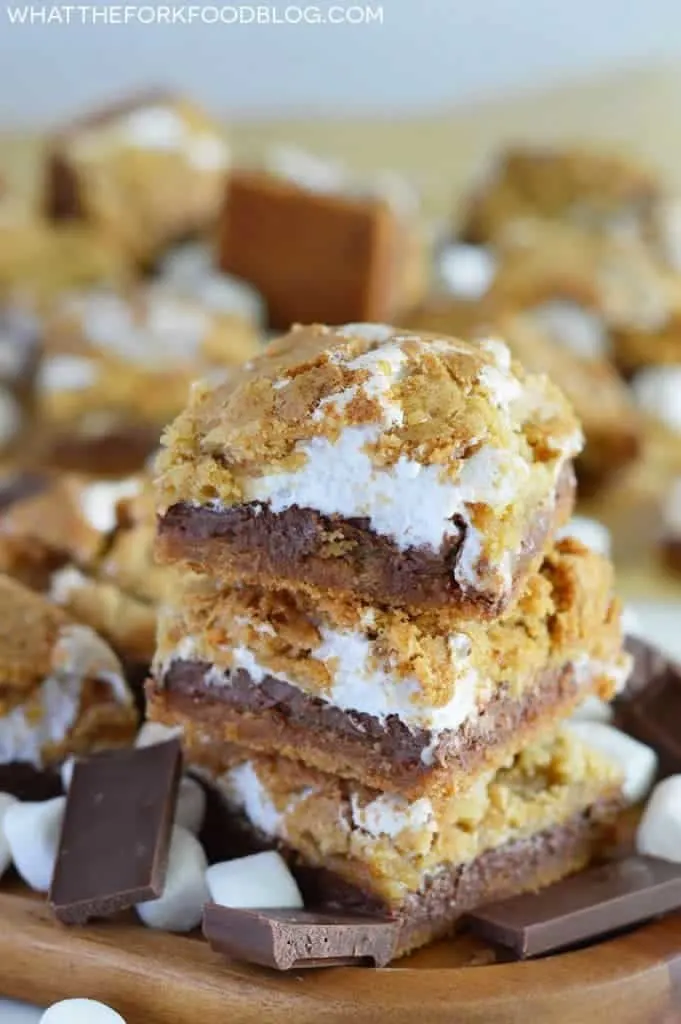 Stacked Gluten Free S'mores Bars on a brown wood platter surrounded by marshmallows and chocolate
