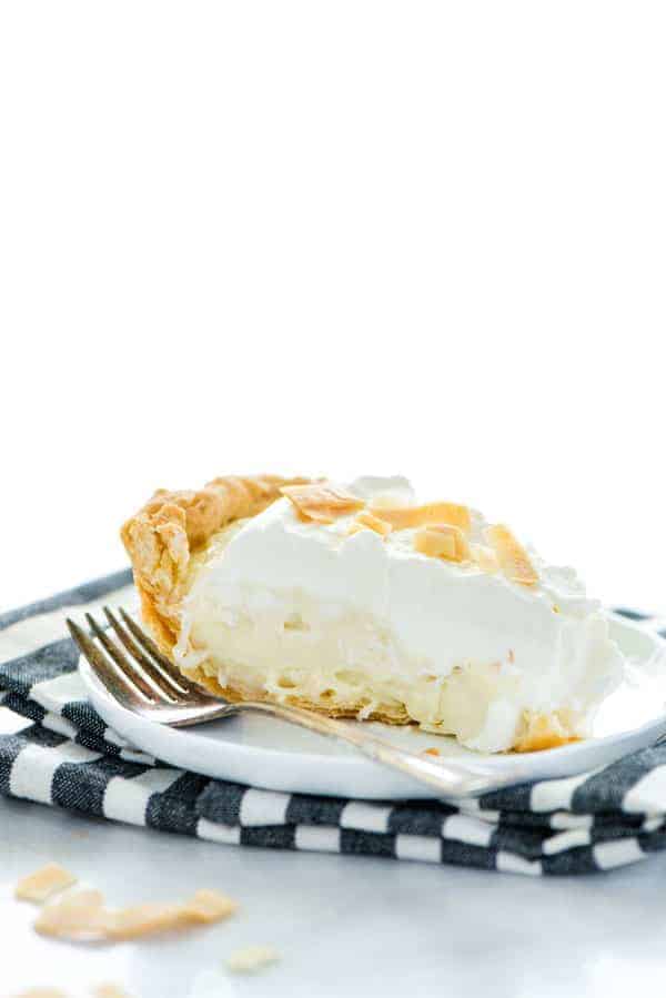 A slice of gluten free coconut cream pie on a white plate with a fork and a black and white striped napkin