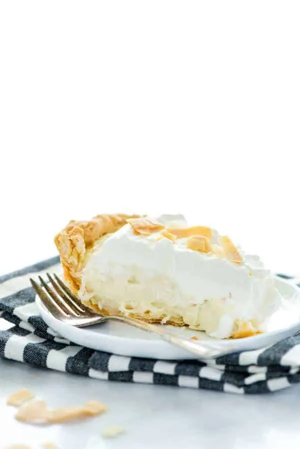 A slice of gluten free coconut cream pie on a white plate with a fork and a black and white striped napkin