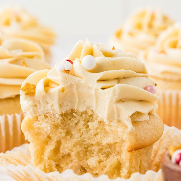Ultra Moist Gluten Free Vanilla Cupcakes frosted with vanilla butter cream frosting. The featured cupcake has the wrapper pealed and a bite taken out.