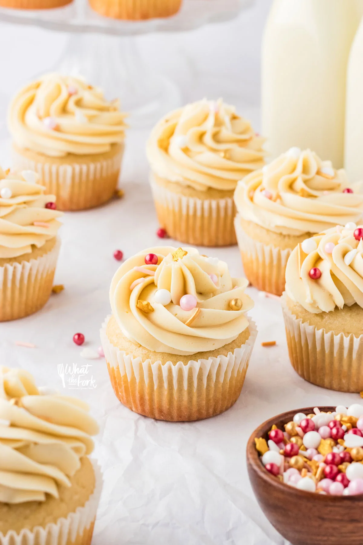 gluten free vanilla cupcakes on a white surface frosted with a swirl of vanilla buttercream frosting and topped with a few red, white, and gold sprinkles