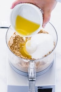 Butter being poured into a food processor with crushed graham cracker crumbs and granulated sugar to show how to make a graham cracker crust