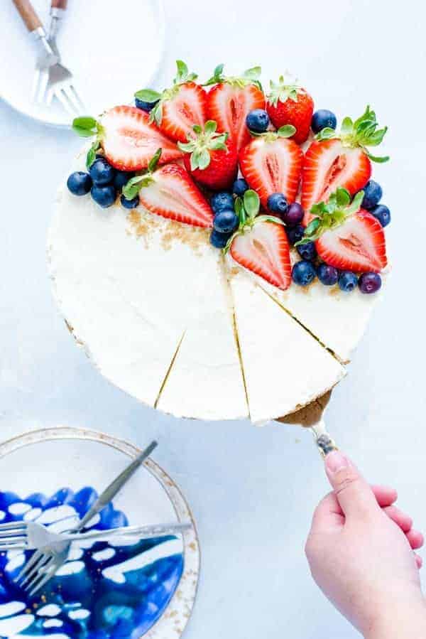Hand using a server to serve a slice of no bake cheesecake topped with fresh strawberries and blueberries