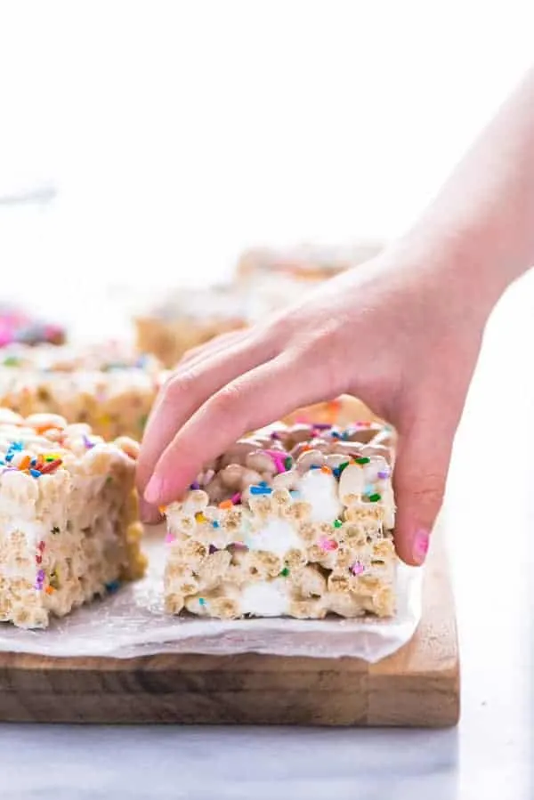 A hand taking a gluten free rice krispies treat off of a brown wood cutting board