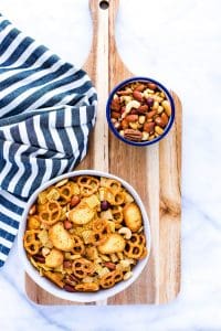 Gluten Free Chex Mix in a white bowl on a wood cutting board with a bowl of mixed nuts and a striped towel.