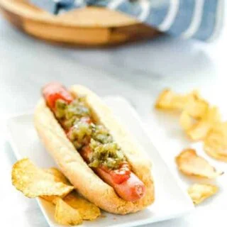 Gluten Free Hot Dog Buns filled with a hot dog and topped with ketchup and relish and potato chips on the side.