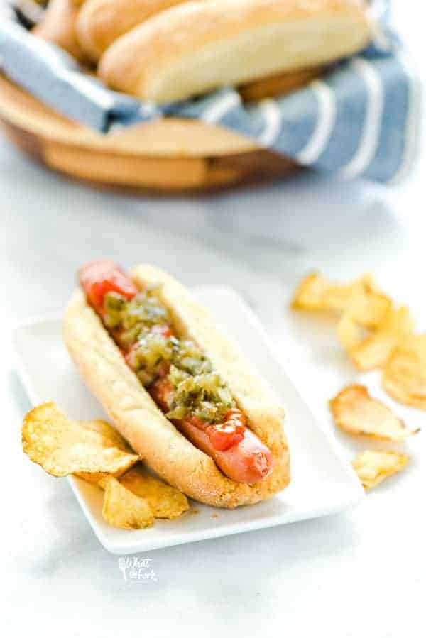 Gluten Free Hot Dog Buns filled with a hot dog and topped with ketchup and relish and potato chips on the side.