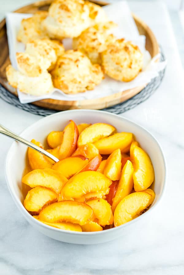 a bowl of sliced fresh peaches with a platter of gluten free drop biscuits in the background to make gluten free peach shortcake