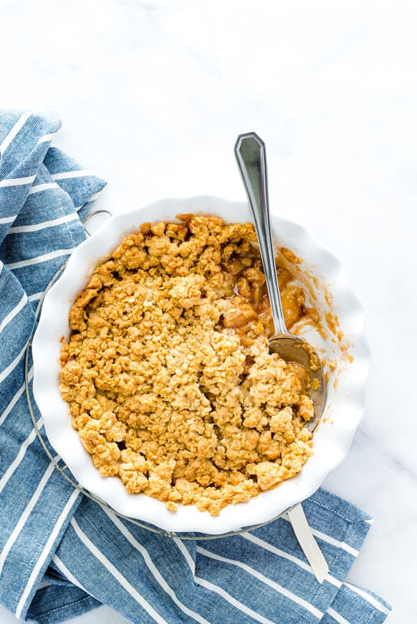GF Apple Crisp in a round white baking dish with a silver spoon on a blue kitchen towel