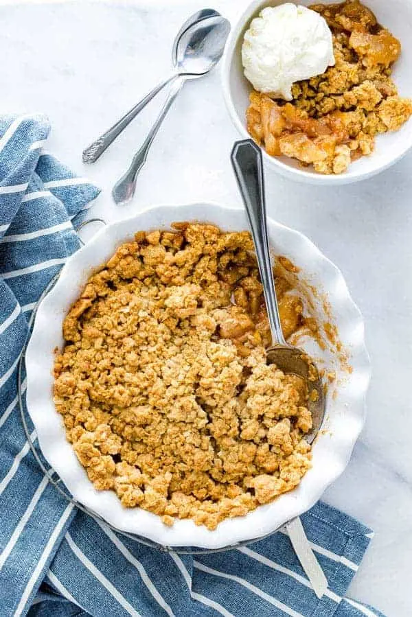 A a dish of apple crisp topped with vanilla ice cream with a plate of a smaller portion on the side.