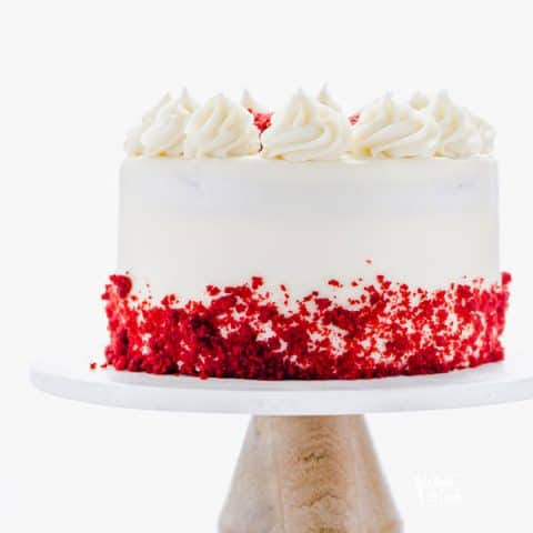 A gluten free Red Velvet Cake on a white marble cake stand with a wood base. The cake is decorated with cream cheese frosting and red velvet cake crumbs.