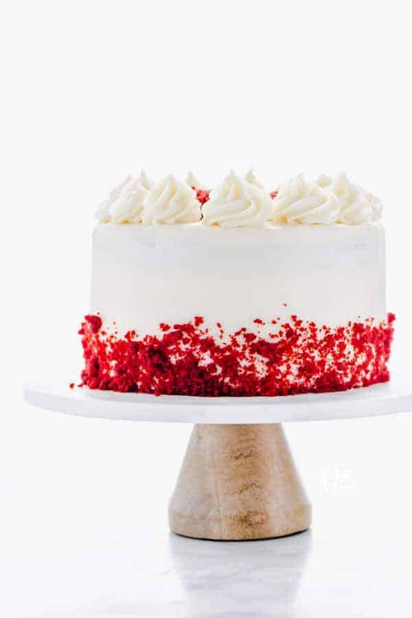 A gluten free Red Velvet Cake on a white marble cake stand with a wood base. The cake is decorated with cream cheese frosting and red velvet cake crumbs.
