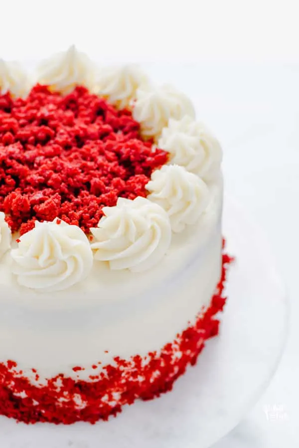 Gluten Free Red Velvet Cake on a white cake stand frosted with cream cheese frosting and decorated with red velvet cake crumbs