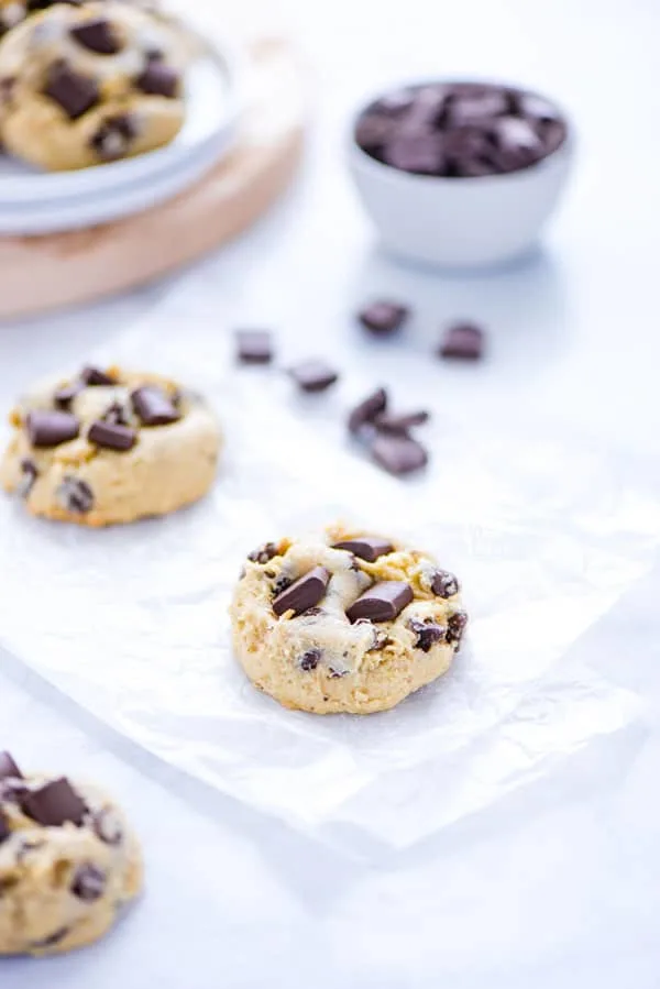 Gluten Free Sourdough Chocolate Chip Cookies on white parchment paper with chocolate chunks and a plate of cookies in the background