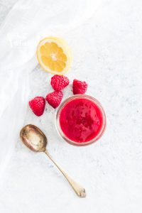 Homemade raspberry sauce in a small glass jar surrounded by fresh raspberries, fresh lemon, and a spoon for scooping