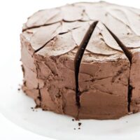 A frosted gluten free sourdough chocolate cake recipe on top of a white cake stand with a wood base.