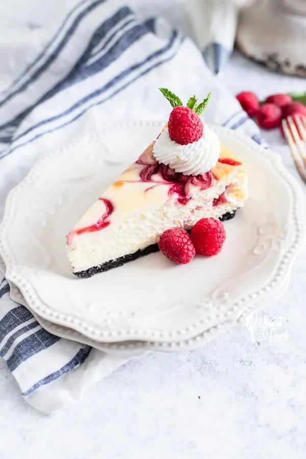 a slice of white chocolate raspberry cheesecake on a white plate garnishes with fresh raspberries, whipped cream, and a sprig of fresh mint