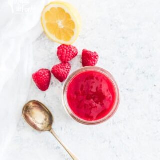 Homemade raspberry sauce in a small glass jar surrounded by fresh raspberries, fresh lemon, and a spoon for scooping