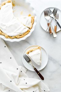 overhead shot of a slice of maple cream pie on a white plate with a wood trimmed fork next to the whole pie and a stack of plates topped with forks