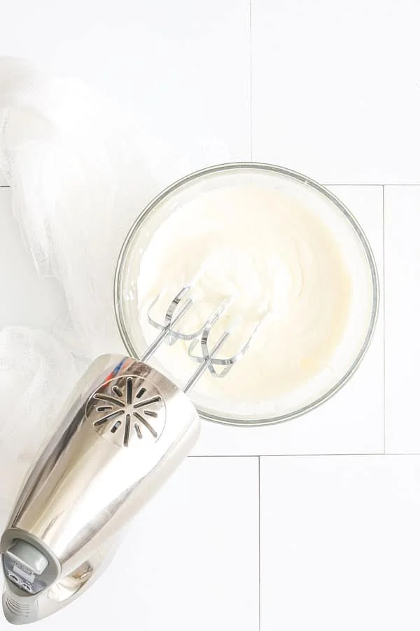 homemade whipped cream being made in a glass bowl with a silver electric hand mixer