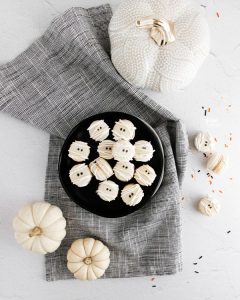Mummy Macarons with Maple Cinnamon Filling on a black plate on top of a black and white cloth napkin surrounded by white pumpkins