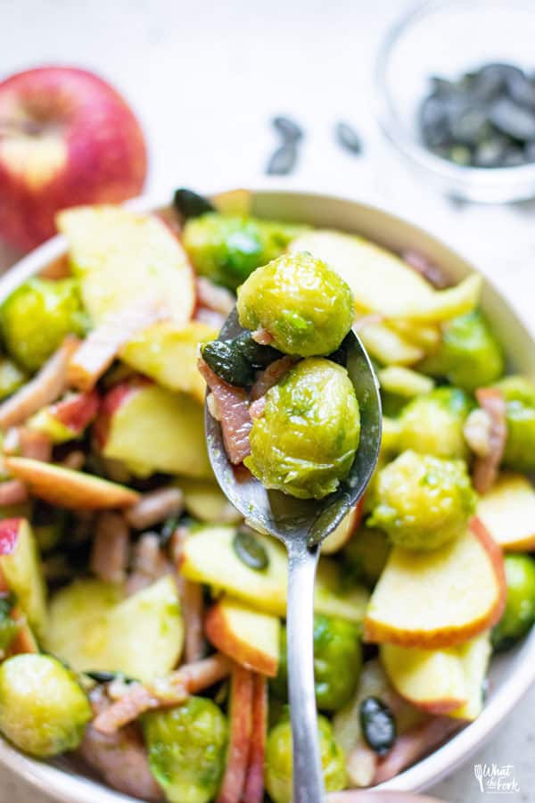 Sautéed Brussels Sprouts with Bacon and Apple in a white serving bowl being dished with a silver serving spoon
