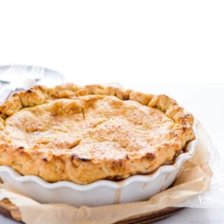 A baked gluten free apple pie in a white pie dish on top of a brown parchment paper and a wood pie board
