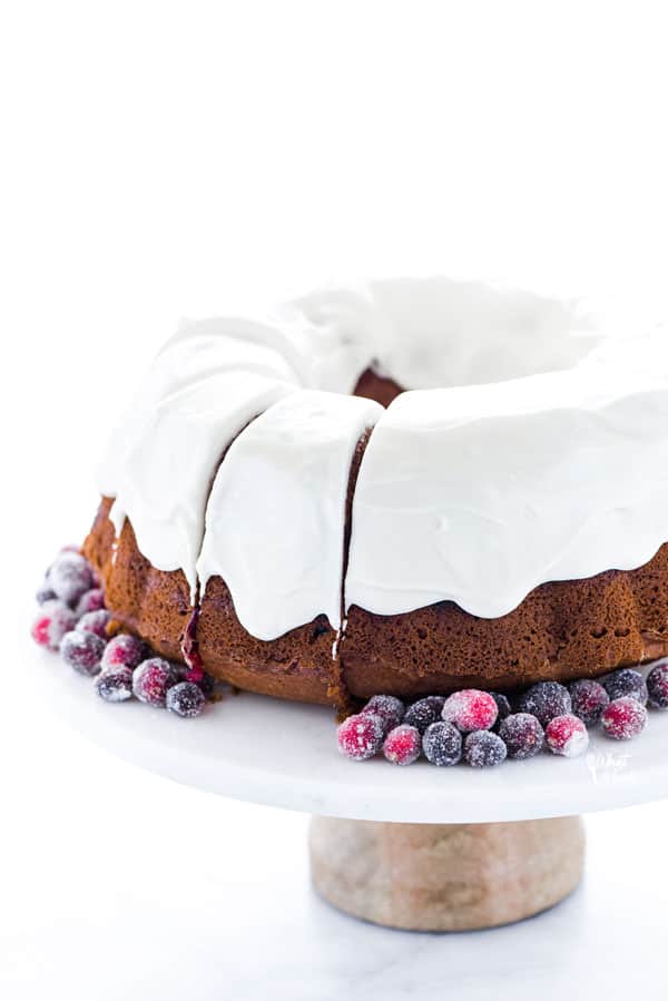 a gluten free cranberry bundt cake with cream cheese icing on a white marble cake stand with wood base garnished with sugared cranberries