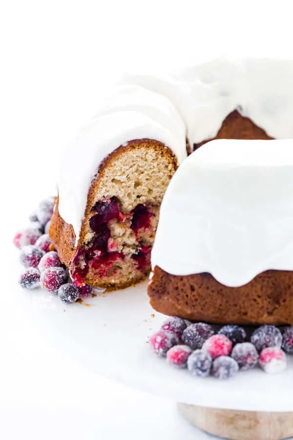 a gluten free cranberry orange bundt cake on a white cake stand garnished with sugared cranberries with a slice removed