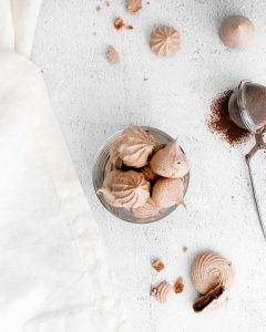overheard shot of dark chocolate meringue cookies in a round class cup on a white backdrop surrounded by a white towel, more chocolate meringue cookies, and a tea ball filled with cocoa powder
