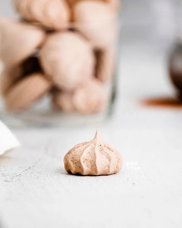 a single dark chocolate meringue cookie on a white surface with a cup full of cookies in the background