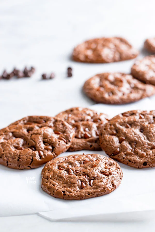 flourless chocolate cookies on a white surface with more cookies and chocolate chips in the background