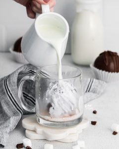 hot milk being poured over a hot chocolate bomb in a clear glass mug
