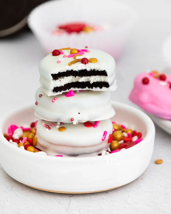 3 Valentine's Chocolate Covered Oreos stacked in a small white bowl filled with sprinkles with a bite mark in the top Oreo