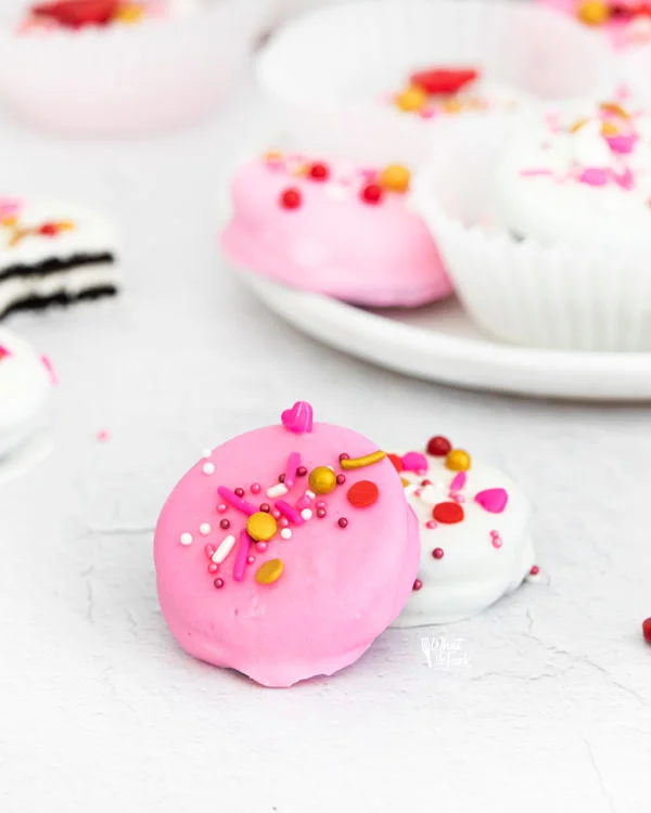 pink and white Valentine's Chocolate Covered Oreos stack on each other on a white surface with more cookies in the background