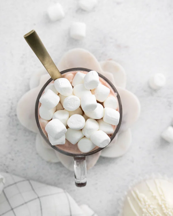 overhead shot of hot cocoa in a clear glass mug with a gold spoon garnished with a pile of mini marshmallows
