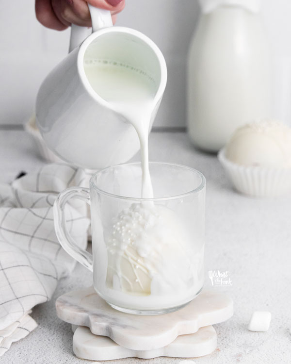a white hot chocolate bomb in a clear glass mug with hot milk being poured over it