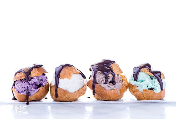 gluten free profiteroles filled with different flavors of ice cream, topped with chocolate ganache, and lined up on a white marble surface
