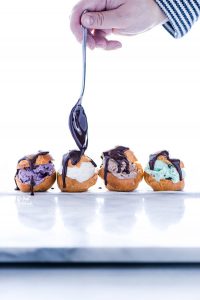 gluten free profiteroles filled with different flavors of ice cream being topped with chocolate ganache