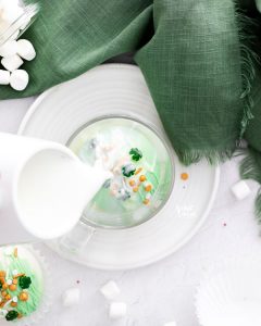 hot milk being poured over a green St. Patrick's Day Hot Chocolate Bomb in a clear glass mug