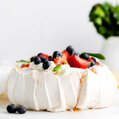 a finished classic pavlova recipe served on a white cake plate and topped with fresh berries and mint