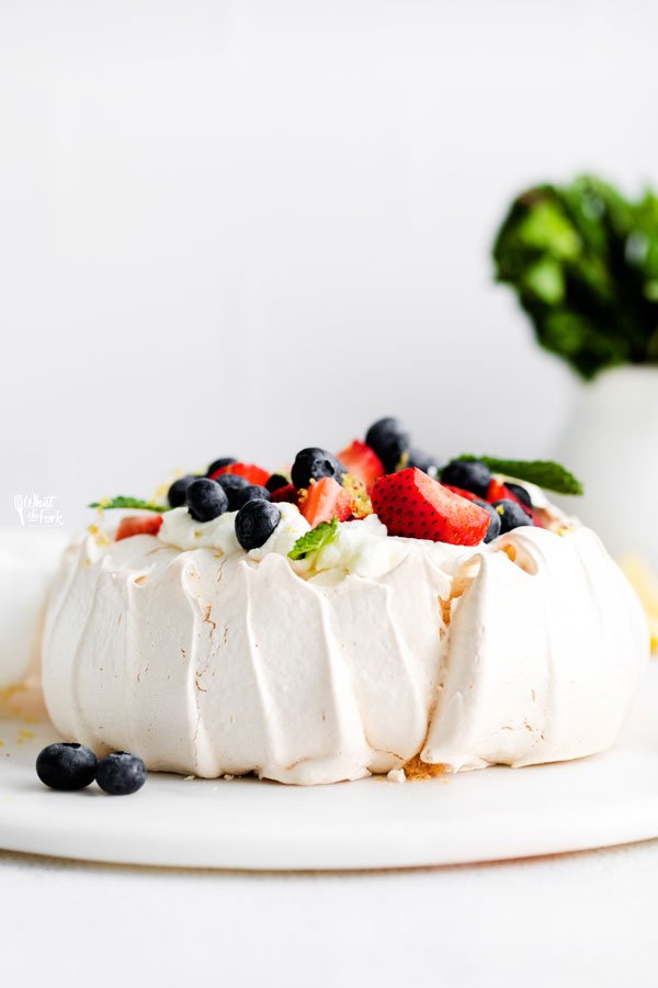 a finished classic pavlova recipe served on a white cake plate and topped with fresh berries and mint