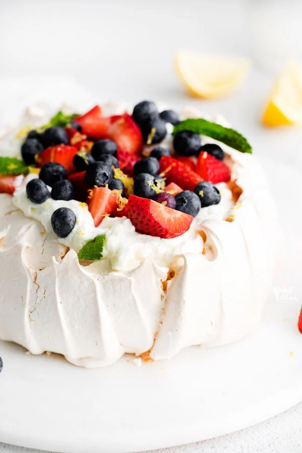 pavlova recipe being served on a flat white cake plate topped with fresh whipped cream, berries, fresh mint, and lemon zest