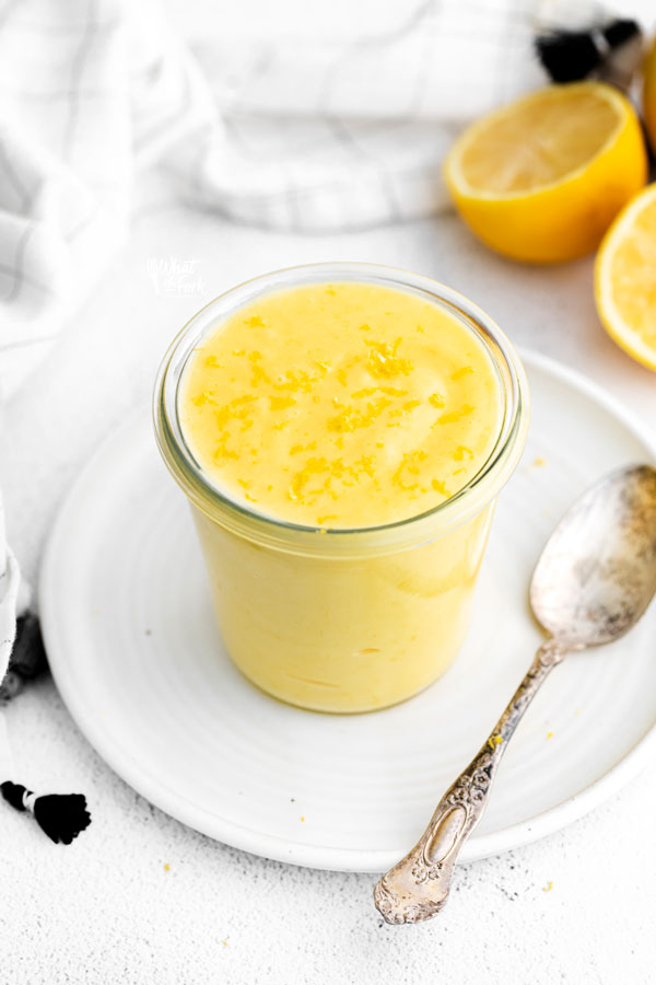 lemon curd topped with lemon zest in a clear glass Weck Jar on top of a white plate with a spoon on the side