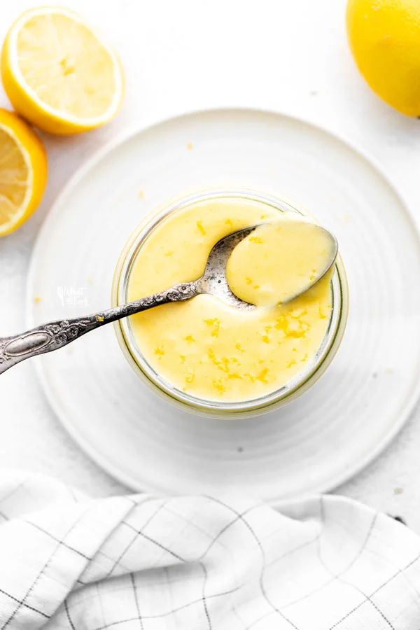 overhead shot of a lemon curd recipe in a glass jar on top of a white plate with a spoonful of curd on top of the jar