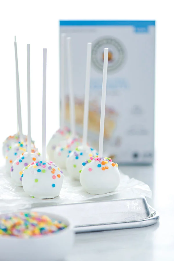 finished Gluten Free Confetti Cake Pop Recipe on an overturned sheet pan lined with wax paper with a box of King Arthur Gluten Free Confetti Cake Mix in the background