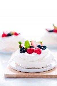 a mini pavlova recipe on a white plate on top of a light wood cutting board. The Mini pavlova is filled with whipped cream and topped with fresh berries and mint