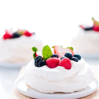 a mini pavlova recipe on a white plate on top of a light wood cutting board. The Mini pavlova is filled with whipped cream and topped with fresh berries and mint