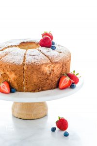 Gluten Free Angel Food Cake on a white and wood cake stand garnished with fresh berries and dusted with powdered sugar