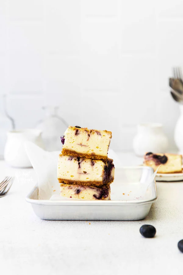 3 Gluten Free Blueberry Cheesecake Bars stacked on a 1/8 sheet pan lined with white parchment paper